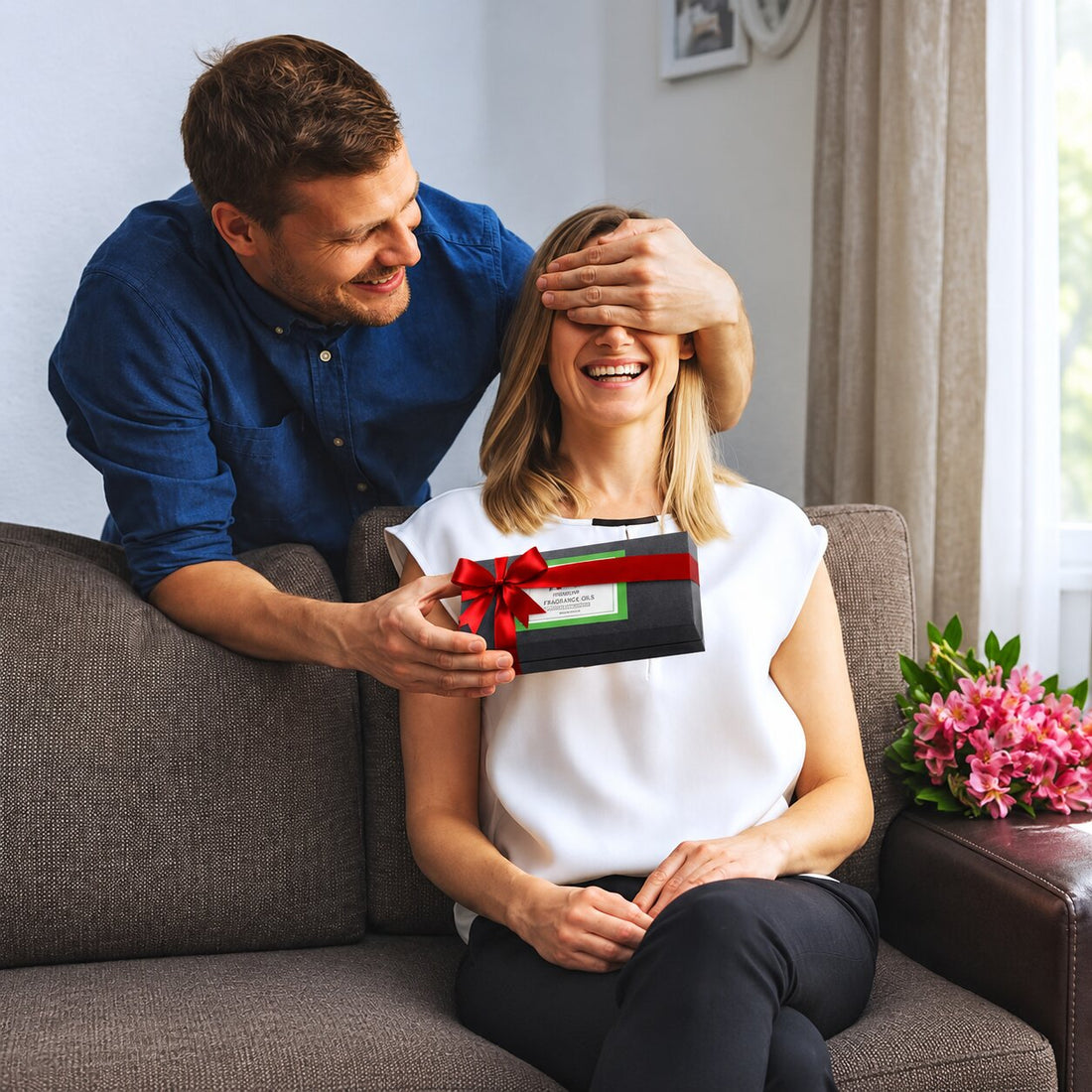 A man in a blue shirt sits on a gray couch behind a woman in a white top, covering her eyes while he holds a My Health A-Z 6 Pack gift box with a red ribbon.