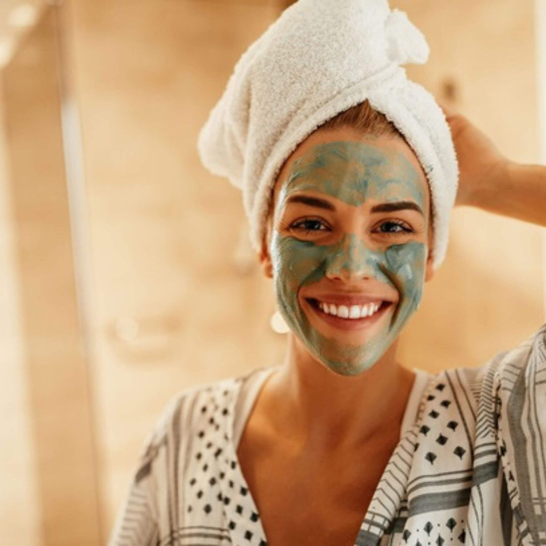Woman with a green facial mask and towel on her head, smiling.