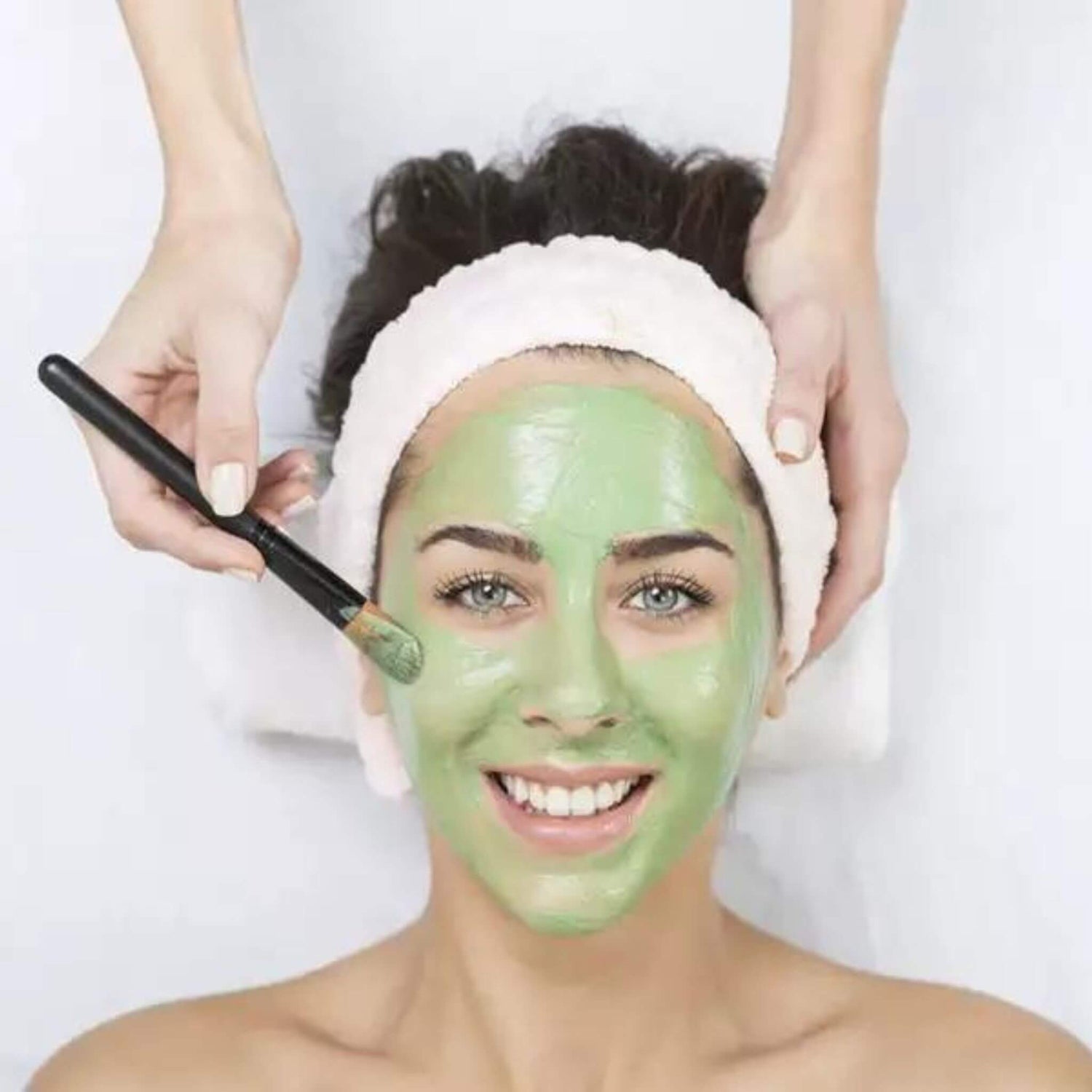 Woman applying green facial mask with a brush on a white background